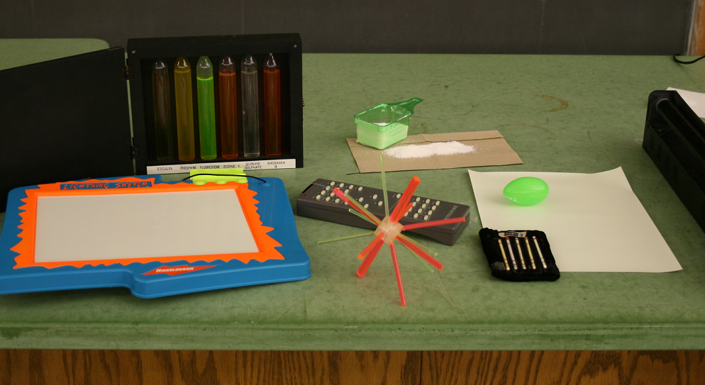 Collection of UV-sensitive objects under normal room lighting: test tubes with colored fluorescent solutions, laundry detergent powder, phosphorescent toys, and other items on a green table