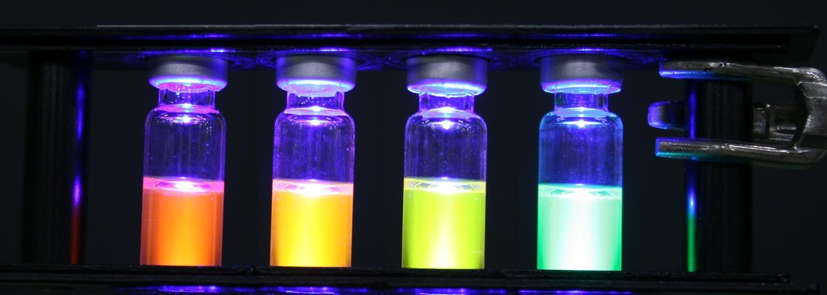 Four sealed vials in a rack illuminated from below by blue LED light, fluorescing orange, yellow, yellow-green, and green from left to right