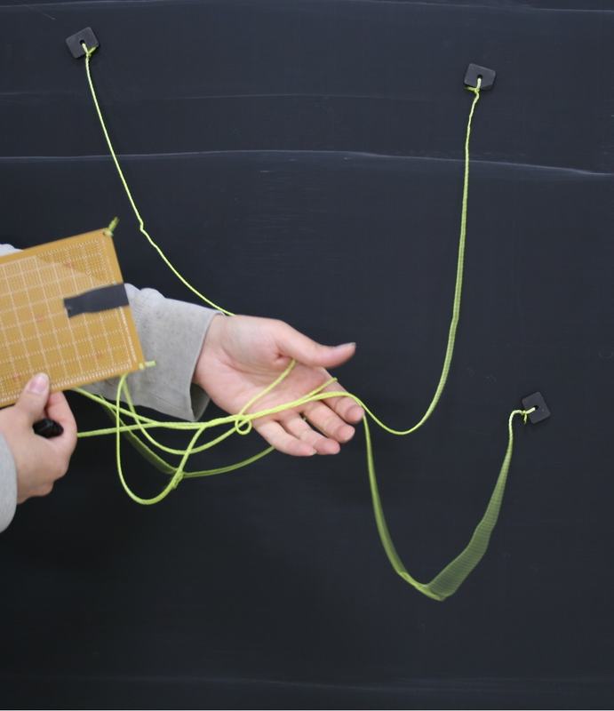 Hand manipulating twisted yellow-green strings with orange board below, strings forming complex loops and crossings