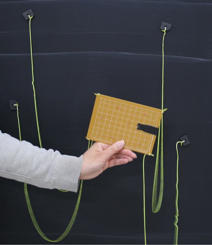 Hand holding rectangular orange perforated board with four yellow-green strings attached to corners, extending to black magnetic anchors on dark surface