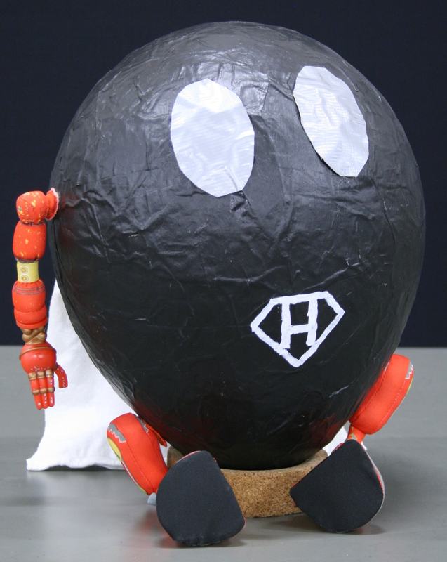 Large black papier-mâché sphere with two white oval eyes and letter H in diamond logo, with orange articulated arms and black feet on cork base