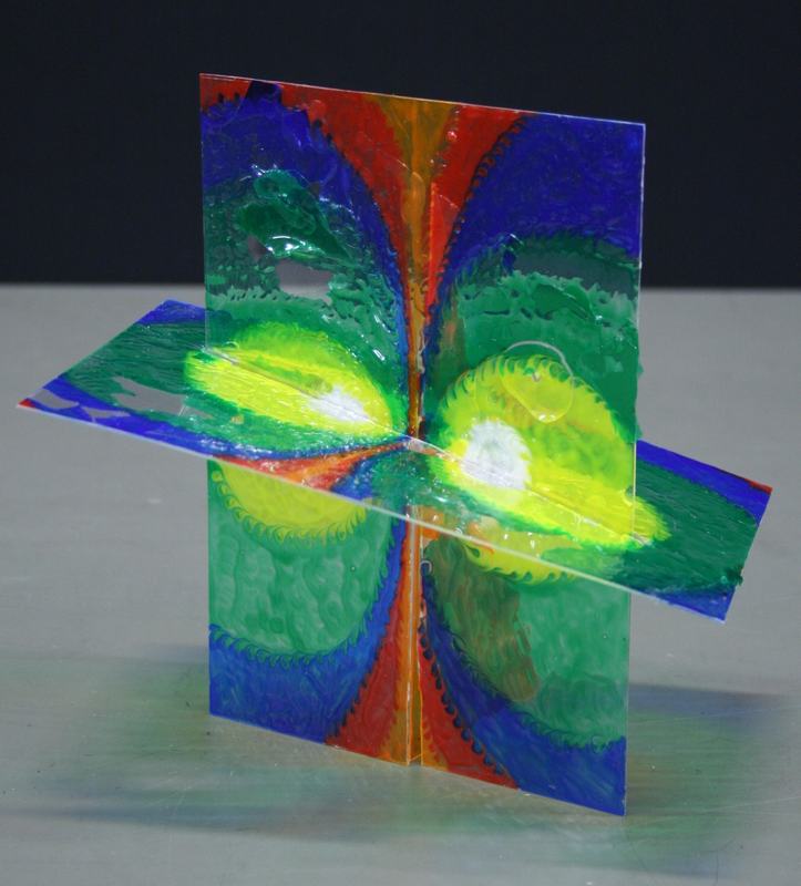 Four transparent plastic sheets arranged in perpendicular cross configuration showing colorful concentric rings in blue, green, yellow, and orange forming clover-like pattern