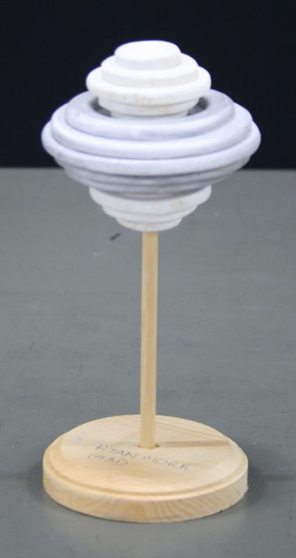 White stacked disc structure resembling spinning top with graduated circular layers decreasing in size toward top and bottom on wooden base