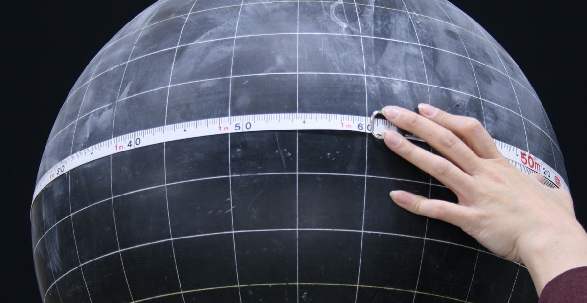 Hand wrapping a flexible measuring tape around the equator of a large dark globe with white grid lines