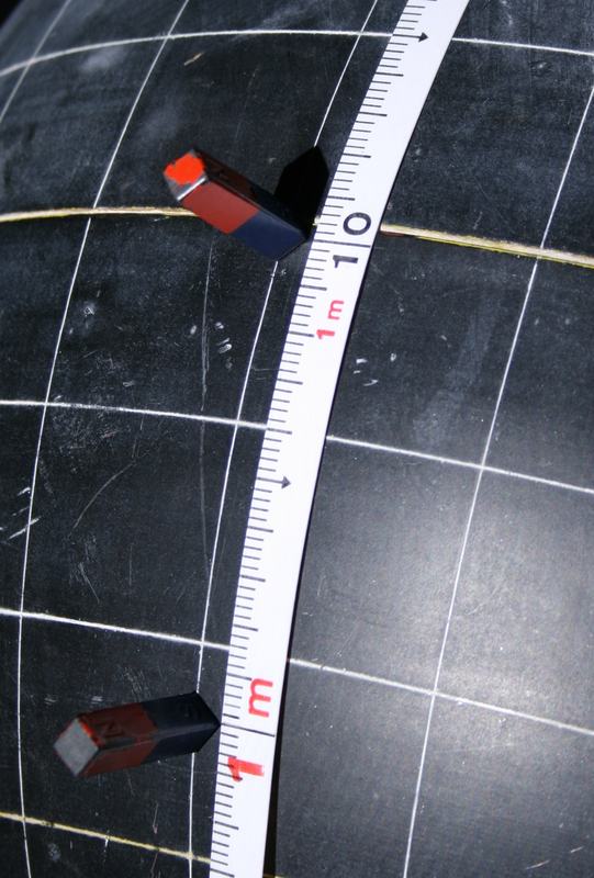 Close-up of a dark globe with white grid lines. Two small red-and-black magnets stand on a meridian, separated by a white measuring tape. The upper magnet casts a visible shadow.