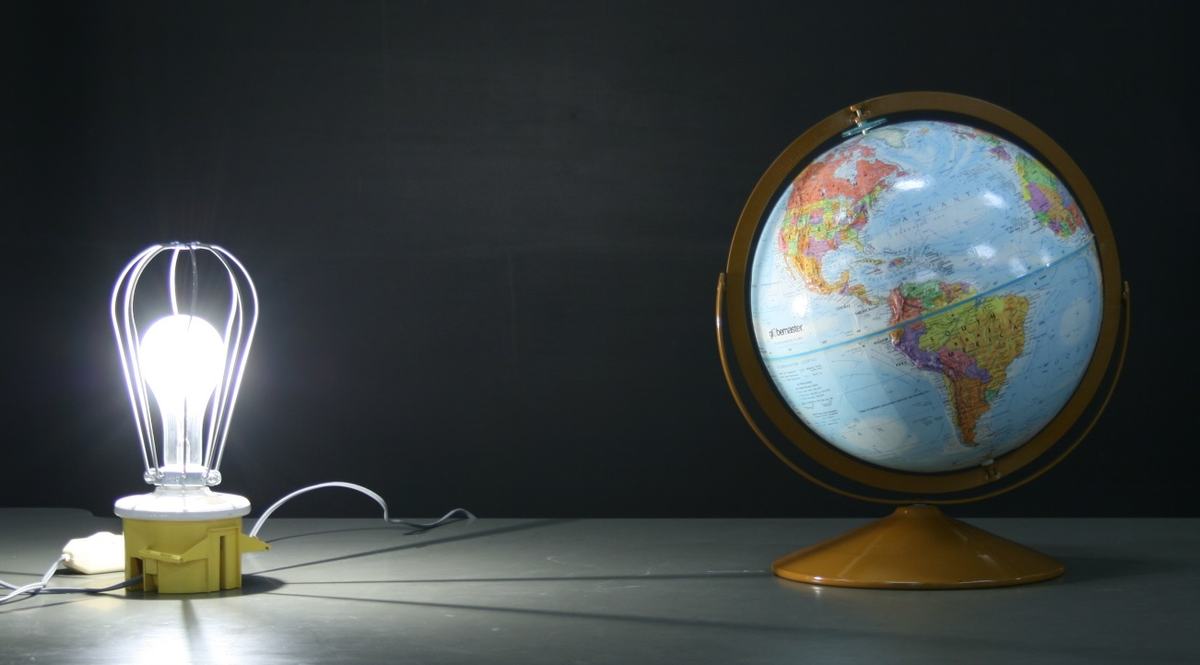 Desktop globe on gold stand next to an illuminated bare bulb in a wire-cage lamp fixture, on a table against a dark background