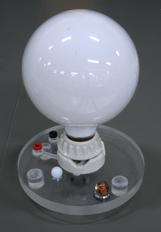 Large white spherical light bulb on a ceramic base mounted on a clear acrylic disc, surrounded by assorted small objects — marbles, peppercorns, small vials, and tiny ball bearings representing planets at scale