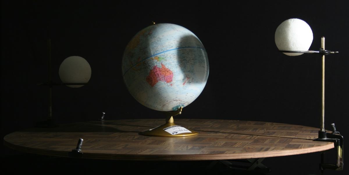 Globe representing Earth on a round wooden table, flanked by two white spheres on clamp stands representing the Moon at different positions, illuminated from one side against a dark background