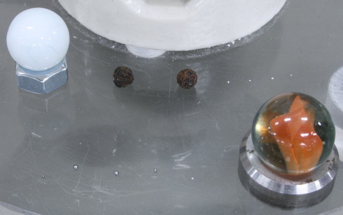Close-up of scale planet models on a clear acrylic surface: a small white marble on a hex nut, two peppercorns, a large decorative marble on a metal ring, and tiny dots barely visible on the surface