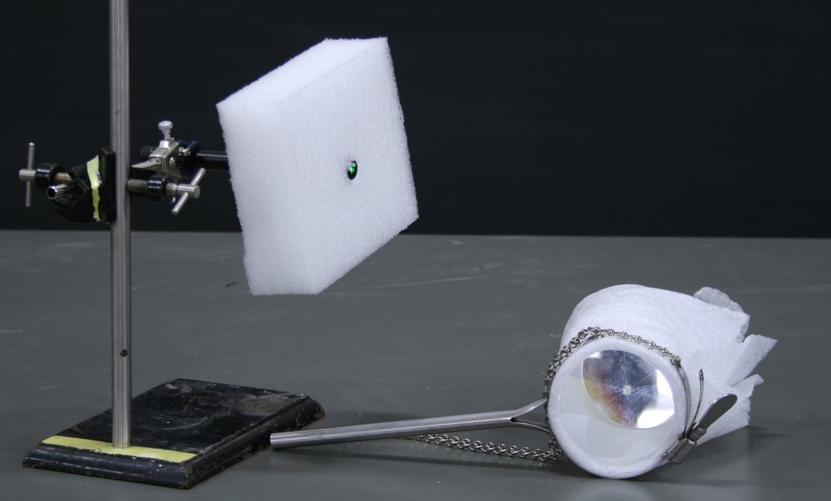 Ring stand with laser pointer clamped to it, aimed at a white foam block target. A corner cube retroreflector wrapped in white foam and secured with a hose clamp on a metal handle lies on the table