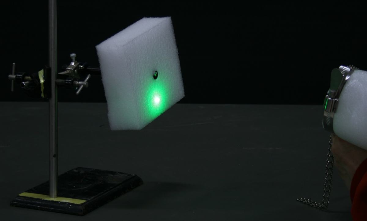 Bright green laser spot on white foam block target, with the foam-wrapped corner cube retroreflector held in the beam path at right