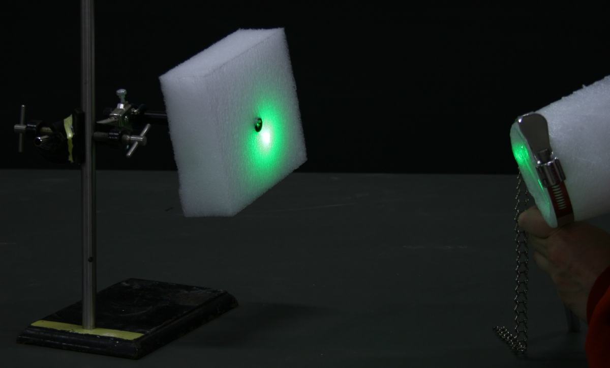 Bright green laser spot on foam block with corner cube held at a different angle, demonstrating that the retroreflected beam returns to the same spot regardless of orientation