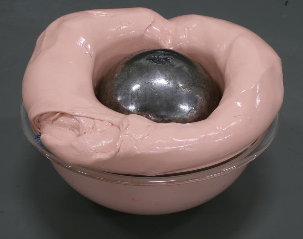 Steel ball nearly engulfed by peach Silly Putty, with only the top of the ball visible above the putty surface