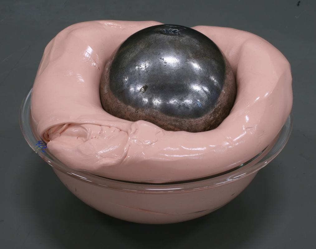 Steel ball partially sunk into peach-colored Silly Putty in a glass bowl, with putty already overflowing the bowl edges