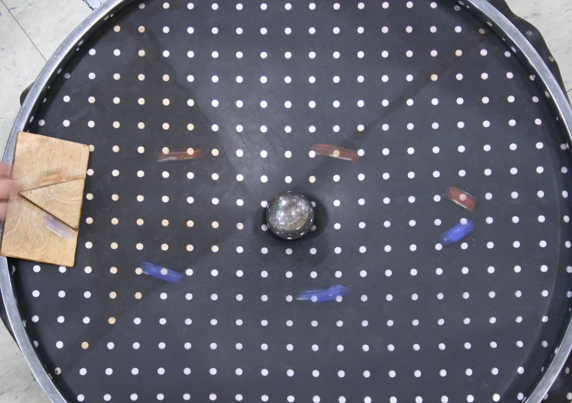 Black spandex sheet with white dot grid stretched between bike wheel rims, with a glass marble at center creating a depression and a wooden triangular launcher at the left edge; colored marbles are scattered on the sheet