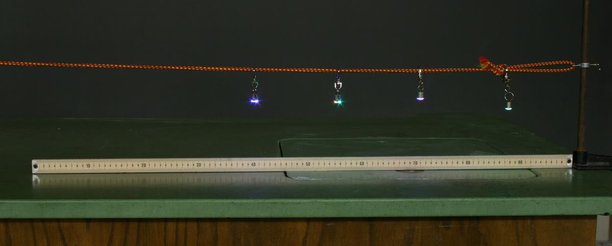 Four flashing LEDs hanging from clips on an orange braided bungee cord at even intervals, positioned above a meter stick on a green lecture bench