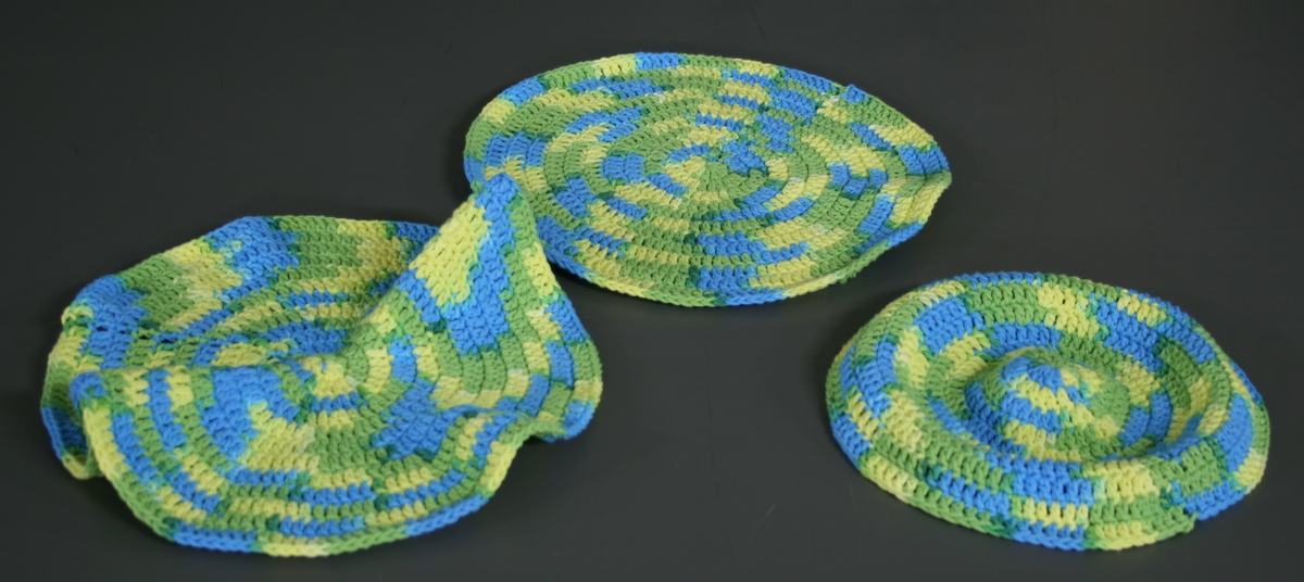 Three crocheted fabric models in variegated blue and green yarn: a flat disk (zero curvature), a ruffled hyperbolic surface (negative curvature), and a dome-shaped spherical cap (positive curvature)